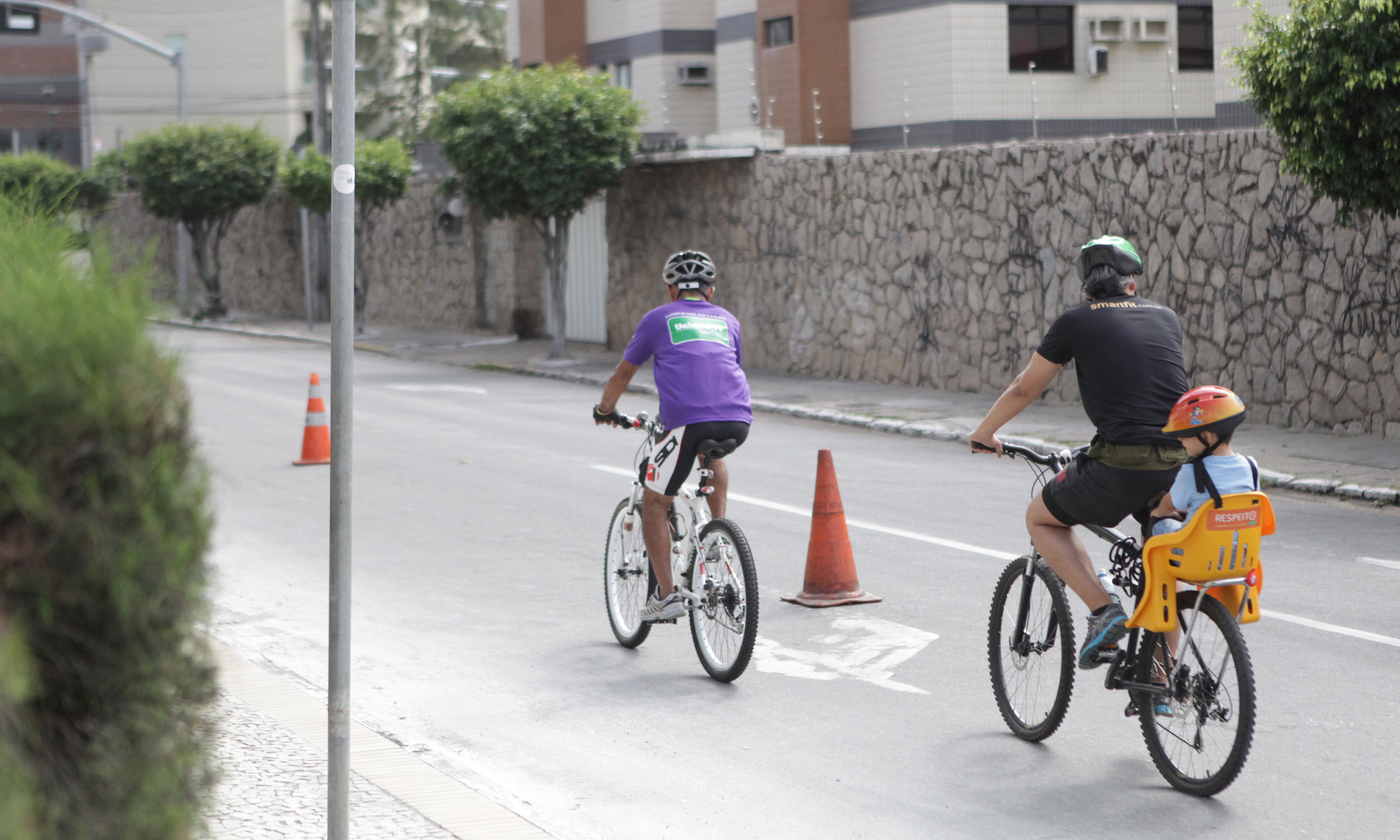 uma mulher e um homem andando de biciciletas. O homem leva uma criança na garupa. Todo os três estão de capacete